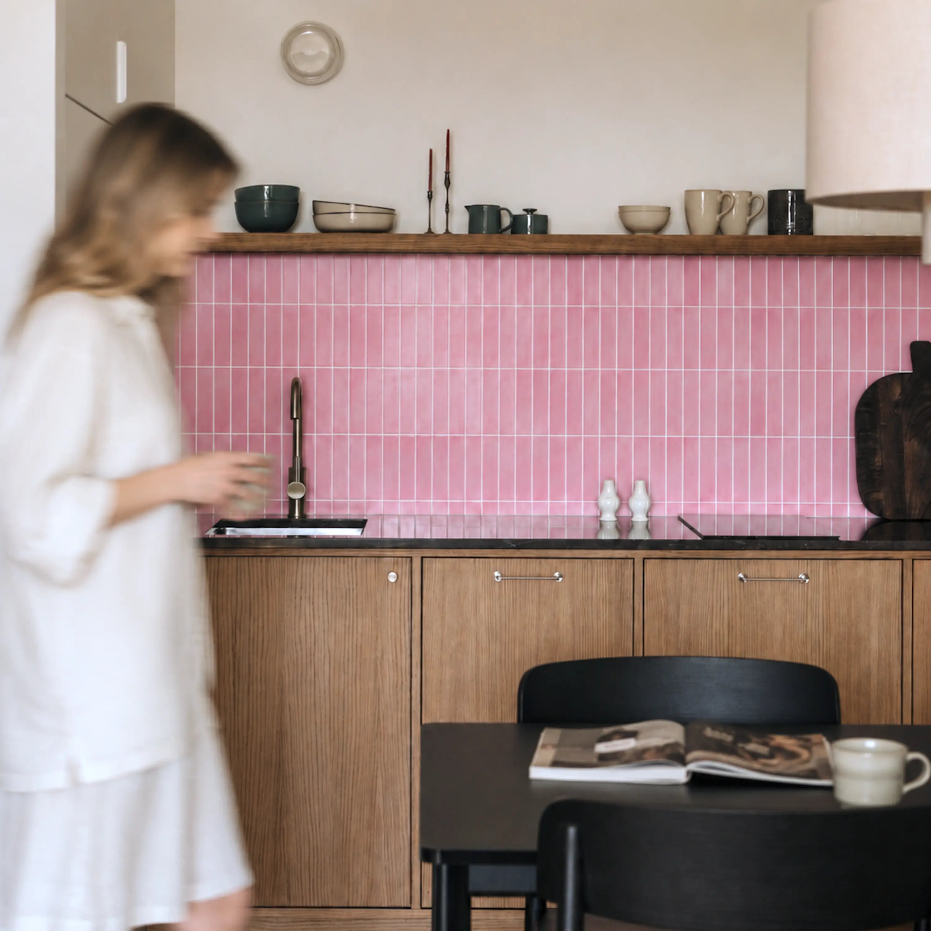 Woman in a warm modern kitchen with Blush Petals pink peel-and-stick backsplash tiles, wood cabinets and a dining area.