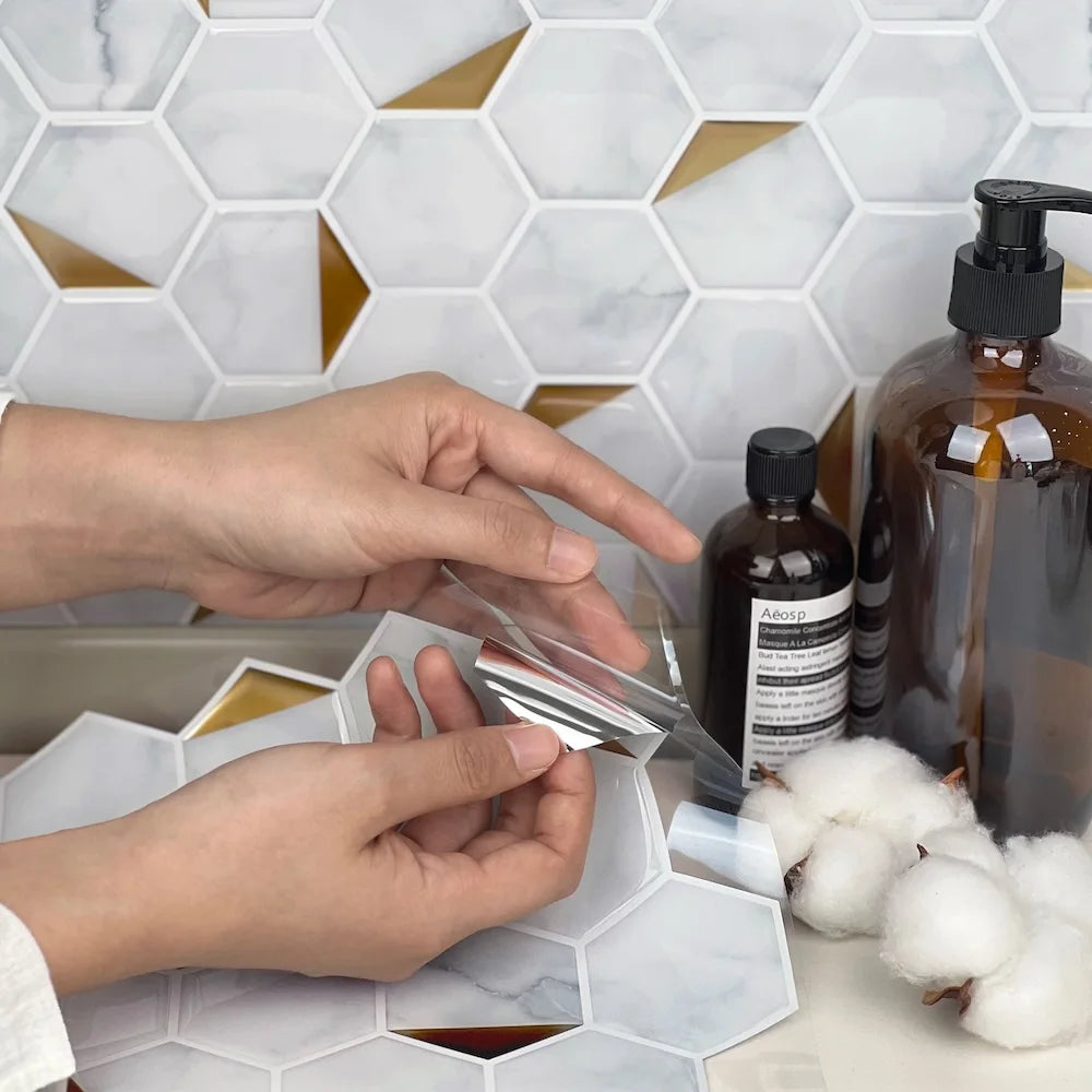 Hands demonstrating the peel-and-stick application of Stickwoll hexagon tiles with marble and gold accents; styled in a bathroom setting with amber bottles and cotton for a luxurious and practical touch