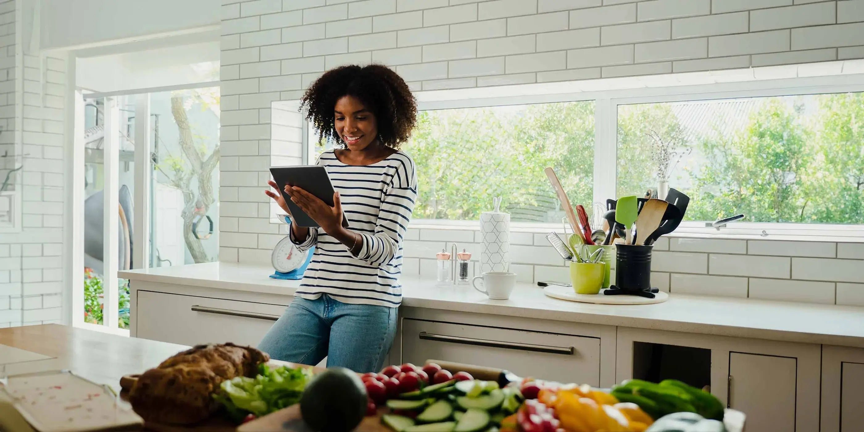 A woman standing in a modern kitchen, smiling while browsing the Stickwoll website on an iPad; the backsplash features Stickwoll hexagon peel and stick tiles, creating a stylish and relaxed atmosphere